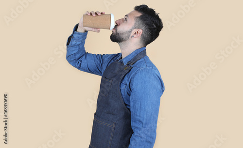 hispanic latino man with apron and shirt stands in profile on yellow background while drinking a coffee