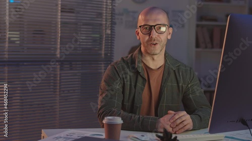 Medium shot of bald mid-adult Caucasian programmer talking at camera sitting at desk in front of computer in dark office at night