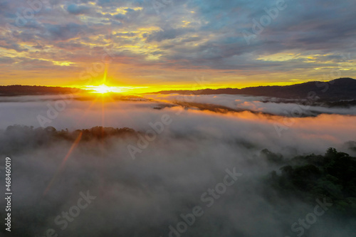 Aerial view during sunrise over a tropical forest with the sun showing up behind the mountains covered in trees and fog with a beautiful cloudscape
