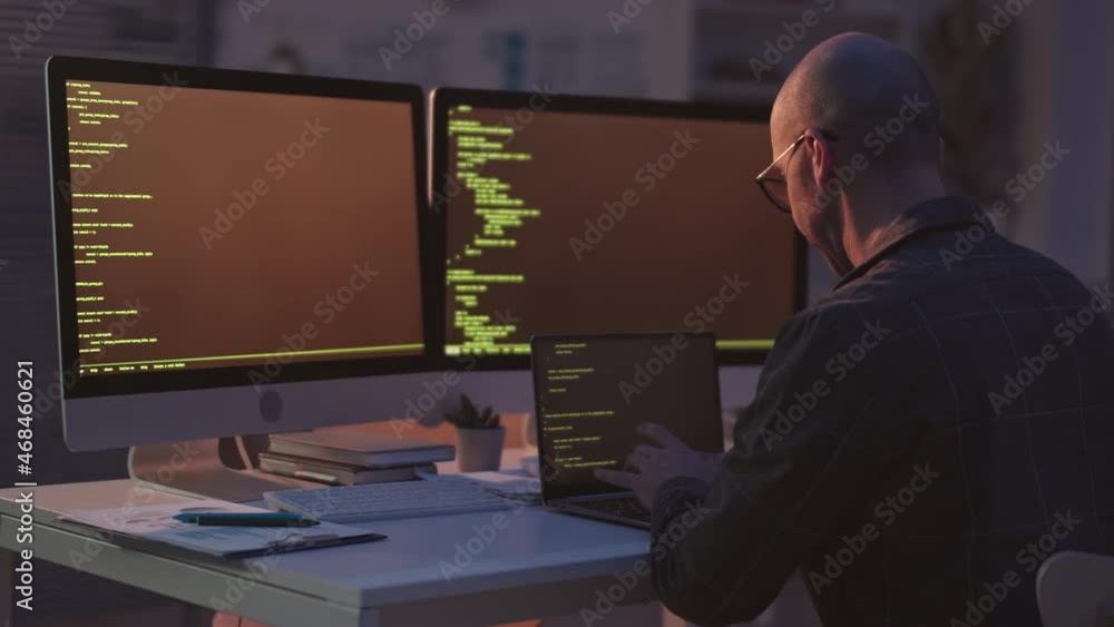 Rear-view medium slowmo shot of bald Caucasian programmer writing program codes on laptop sitting at desk with two monitors in office at night Rear-view medium slowmo shot of bald Caucasian programmer writing program codes on laptop sitting at desk with two monitors in office at night