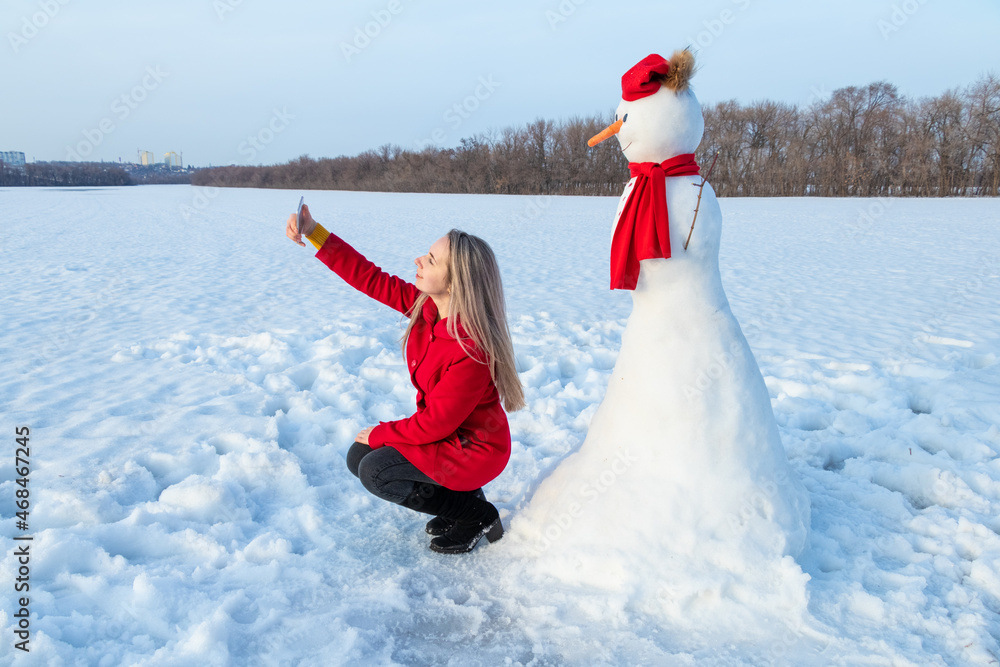 Obraz premium Attractive woman in red coat taking selfie with snowman using smartphone