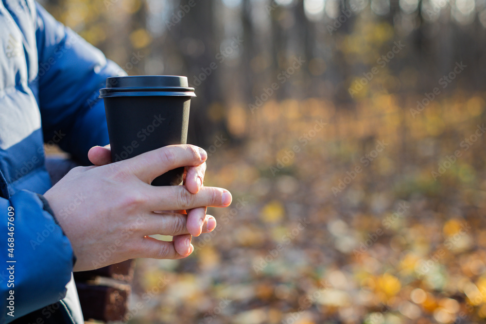 A man holds a disposable paper cup with a drink in the park in the fall. The concept of drinks, tea, coffee, autumn, warm up, rest. A man holds a disposable paper cup with a drink in the park in the fall. The concept of drinks, tea, coffee, autumn, warm up, rest.