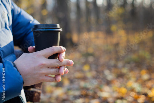 A man holds a disposable paper cup with a drink in the park in the fall. The concept of drinks, tea, coffee, autumn, warm up, rest.