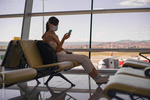 Woman in face mask waiting boarding in big empty airport. Female traveler look at phone, search for boarding pass or flight cancelation information.
