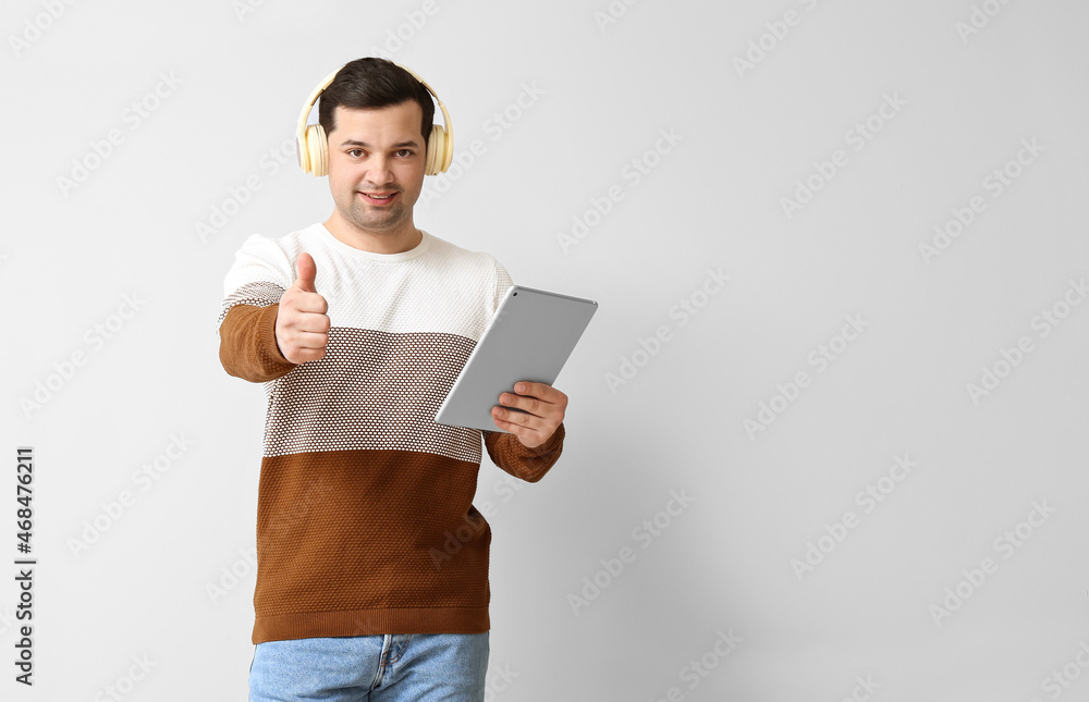 Young man with tablet computer and headphones showing thumb-up on light background Young man with tablet computer and headphones showing thumb-up on light background