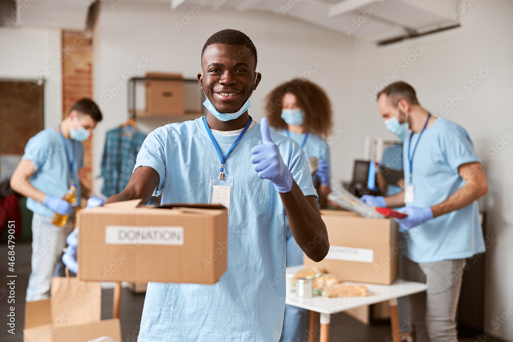 African american young male volunteer in blue uniform, protective mask and gloves smiling, showing thumbs up and holding cardboard box for Donation African american young male volunteer in blue uniform, protective mask and gloves smiling, showing thumbs up and holding cardboard box for Donation