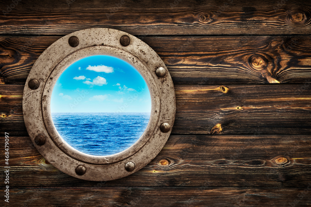 Fototapeta premium Close-up of an old rusty closed porthole window with ocean and blue sky view. Old rich wood grain texture background with knots.