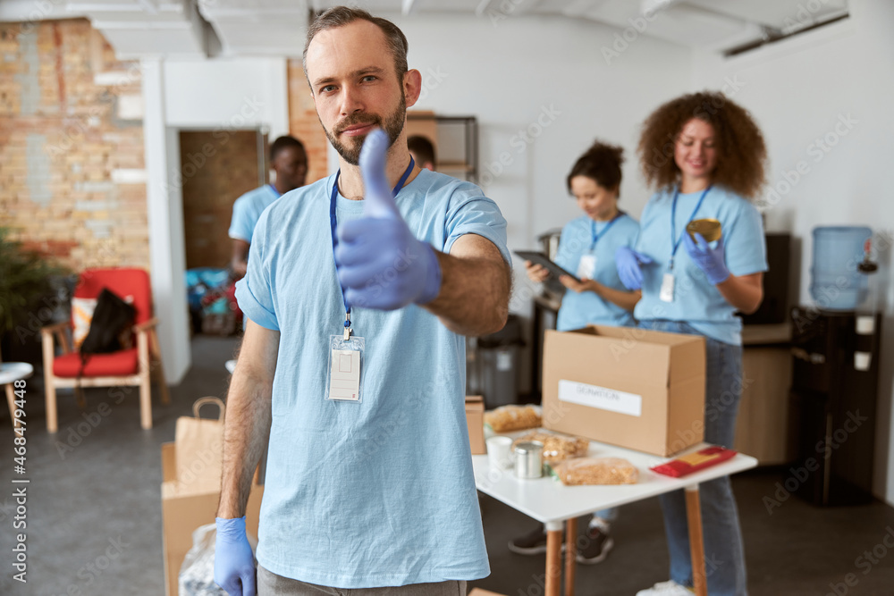 Obraz premium Portrait of caucasian male volunteer in blue uniform and protective gloves showing thumbs up, looking at camera. Team sorting, packing items in cardboard boxes