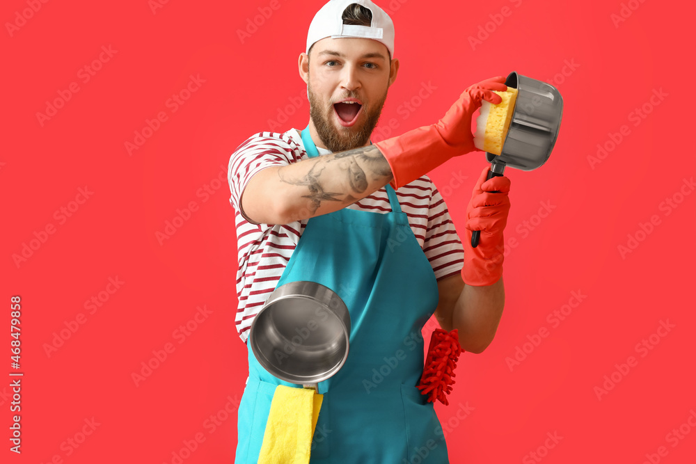 Handsome young man with sponge cleaning cooking pot on red background
