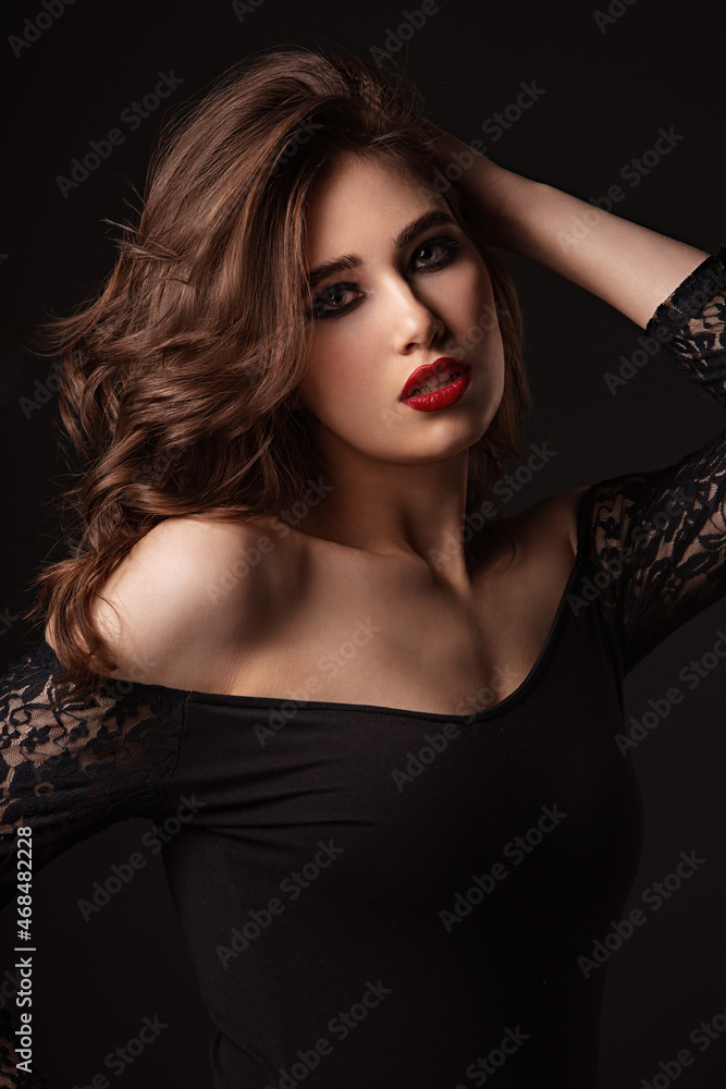 Beautiful bright makeup woman with long brown curly volume hair style, red lipstick, and black smokey eyes looking vamp and passion on black background. Closeup