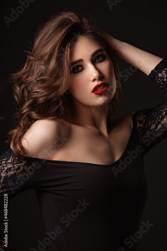 Beautiful bright makeup woman with long brown curly volume hair style, red lipstick, and black smokey eyes looking vamp and passion on black background. Closeup