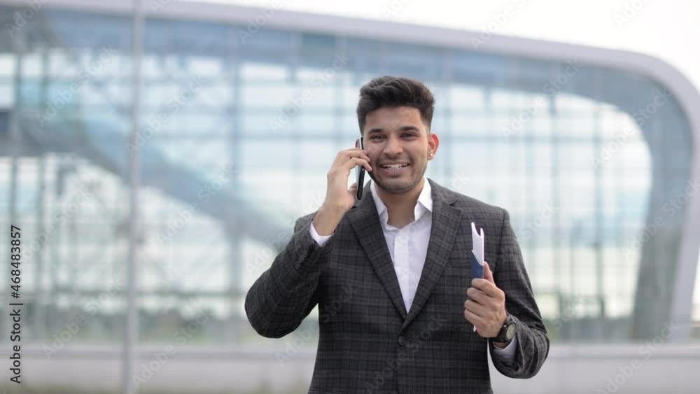 Business, people, communication and travel concept. Close up of positive Arabian businessman, talking on phone and holding passport and ticket, standing outside of modern airport