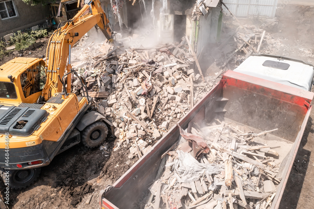 Fototapeta premium Excavator loads construction waste into truck. Demolition of dilapidated housing.