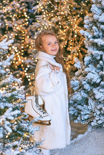 cute caucasian blonde girl in white eco fur coat walking with skates in the winter christmas forest. New year, fairy tale, sports, vacation, holidays, fashion concept