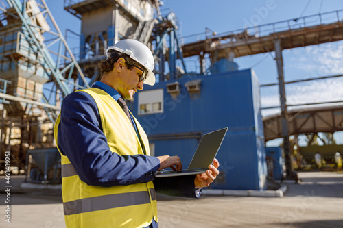 Matured man in work vest standing on territory of manufacturing plant and using laptop