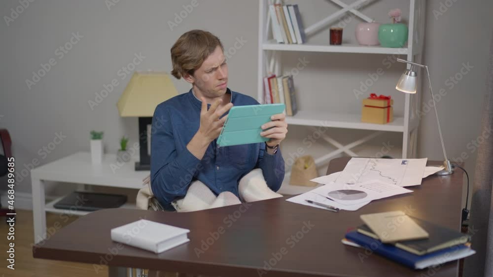 Intelligent disabled young man in wheelchair messaging online on tablet sitting in home office indoors. Portrait of confident Caucasian positive freelancer texting e-mail smiling in slow motion Intelligent disabled young man in wheelchair messaging online on tablet sitting in home office indoors. Portrait of confident Caucasian positive freelancer texting e-mail smiling in slow motion