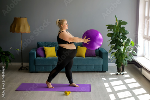 A fat, obese pregnant lady is doing Pilates at home, lifting a fit ball above herself. Home workouts in Interior room with a large window, sofa and yellow-purple pillows. Weight loss in quarantine 