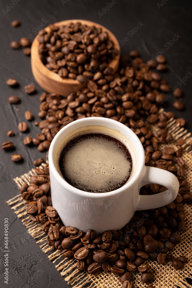 Coffee beans scattered on a rustic black table and white lungo cup with hot coffee drink. Close up image Coffee beans scattered on a rustic black table and white lungo cup with hot coffee drink. Close up image