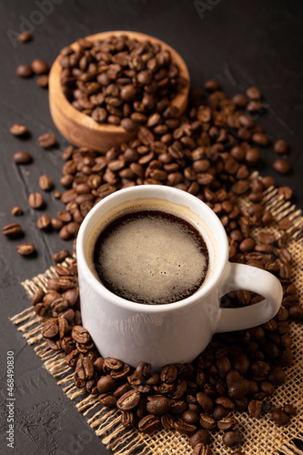 Coffee beans scattered on a rustic black table and white lungo cup with hot coffee drink. Close up image