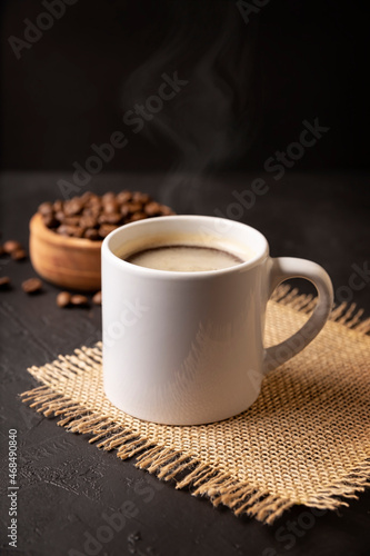 White lungo cup with hot coffee drink and toasted coffee beans scattered on a rustic black table. Close up image. Blank cup for copy space