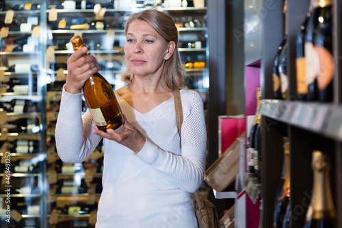 Smiling middle aged female looking for perfect wine for solemn occasion in wine store