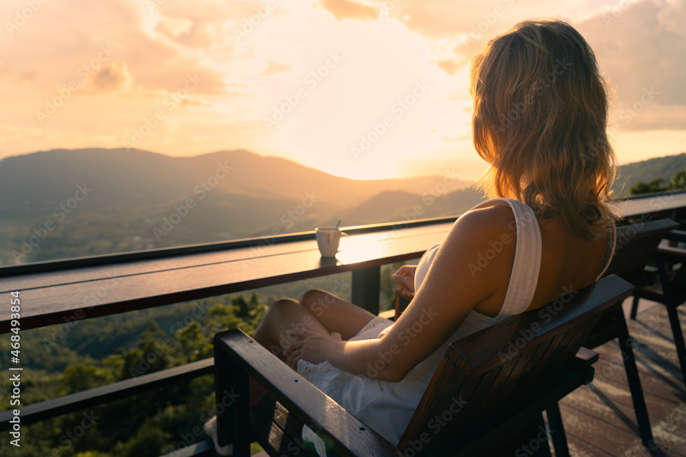 Young slender woman in a white dress is sitting alone with a cup of coffee in a cafe on a hight view point and enjoying the colorful sunset over the valley. In the background sea and mountains.