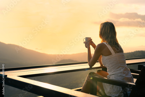 Young slender woman in a white dress is sitting alone with a cup of coffee in a cafe on a hight view point and enjoying the colorful sunset over the valley. In the background sea and mountains.