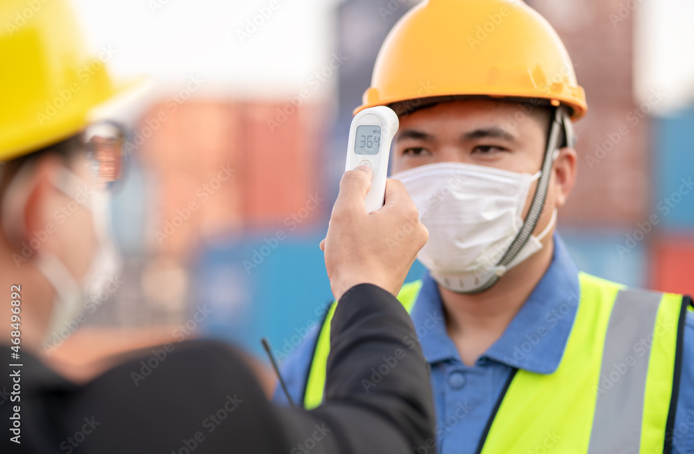 Men at work wearing face mask prevent Covid-19 infection.Manager or engineer foreman using infrared thermometer measure temperature to construction site staff worker at shipping container warehouse. Men at work wearing face mask prevent Covid-19 infection.Manager or engineer foreman using infrared thermometer measure temperature to construction site staff worker at shipping container warehouse.