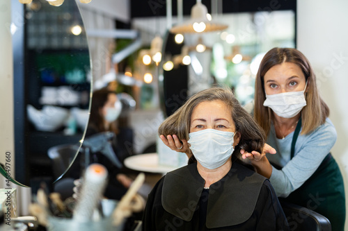 Elderly woman wearing protective face mask sitting in chair in hair salon while professional stylist proposing new hairstyle. New life reality during COVID 19 pandemic