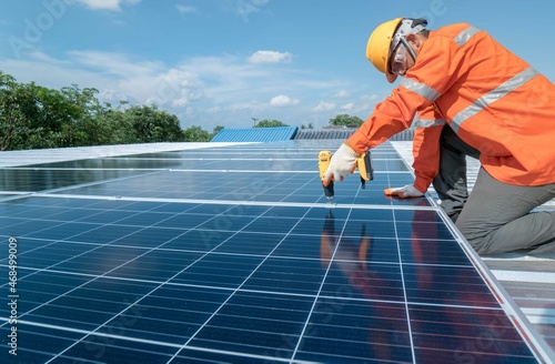 Technician with drill installing solar panels on roof.Male in protective helmet installing solar photovoltaic panel using screwdriver mounting solar module on roof. Alternative energy ecological.