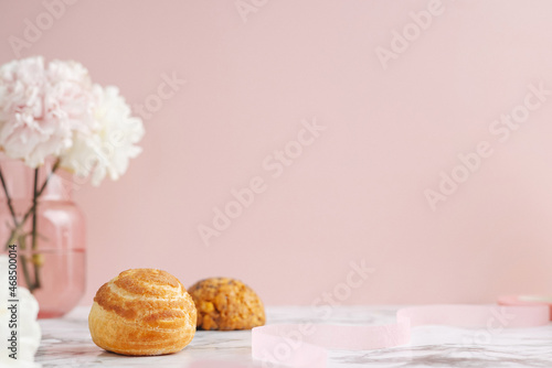 Homemade profitroles pastry filled with custard. Eclairs with cream, French dessert and flowers on marble table on pink background 
