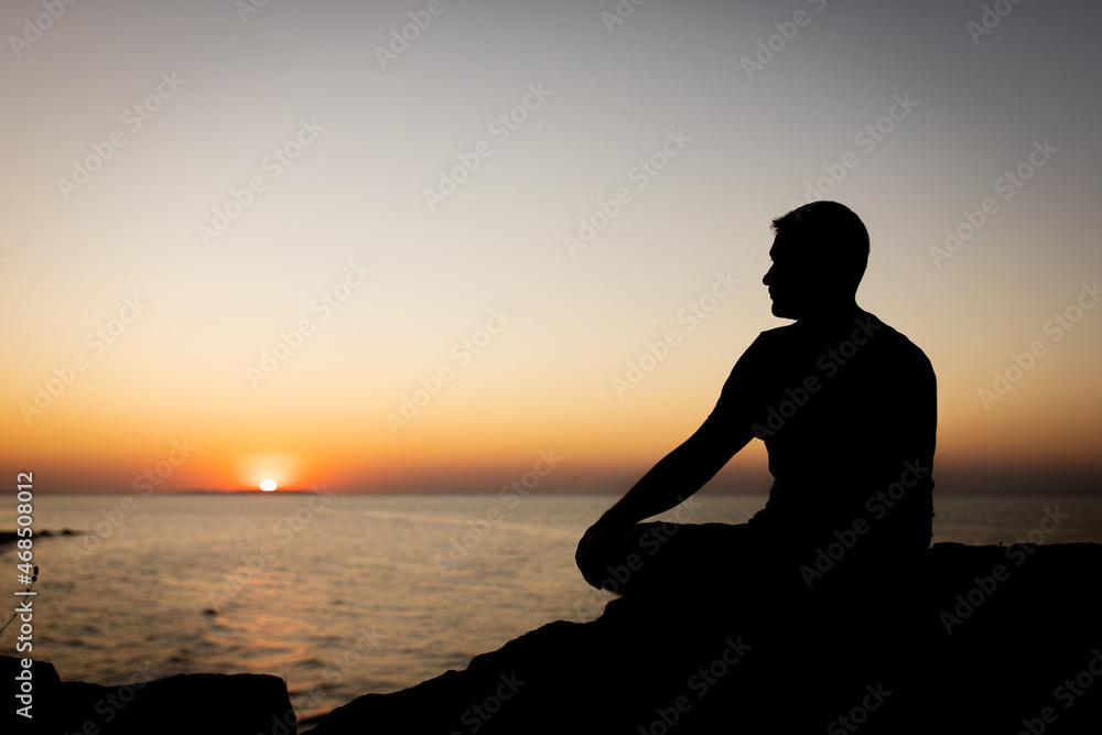 A man enjoys the view of the sunset on the sea, sitting on a rock. Side view, silhouette.