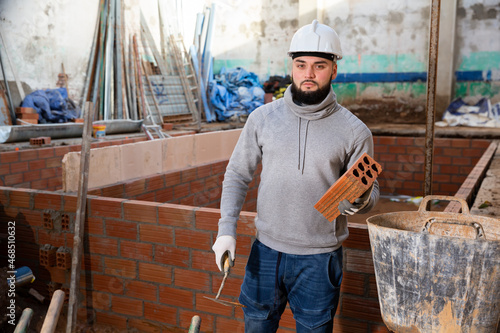 Bilde på lerret Young bearded bricklayer installing brick wall in building under construction