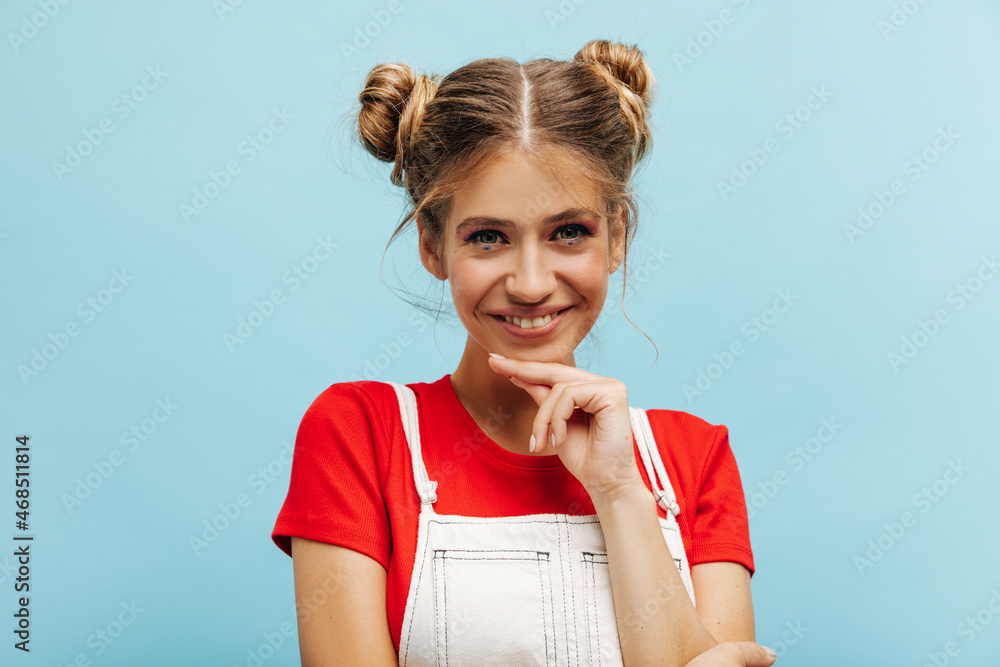 Close-up of stylish young caucasian woman looking at camera while holding her chin over blue background. Smiling girl with two bundles of hair, dressed in casual clothes. Close-up of stylish young caucasian woman looking at camera while holding her chin over blue background. Smiling girl with two bundles of hair, dressed in casual clothes.