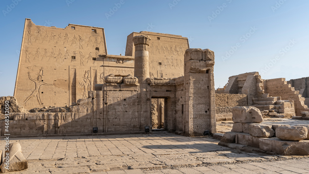 The Temple of the Horus in Edfu. Against the background of the blue sky, high walls with carvings, a column, ancient stones are visible. Egypt