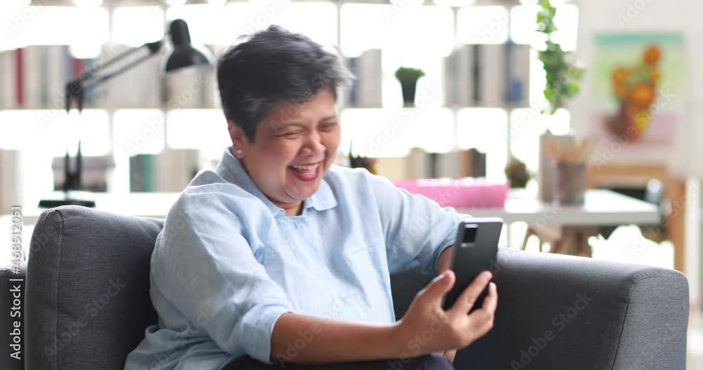 Happy Asian woman laughing at funny joke while sitting on sofa and making video call via cellphone on weekend day at home Happy Asian woman laughing at funny joke while sitting on sofa and making video call via cellphone on weekend day at home