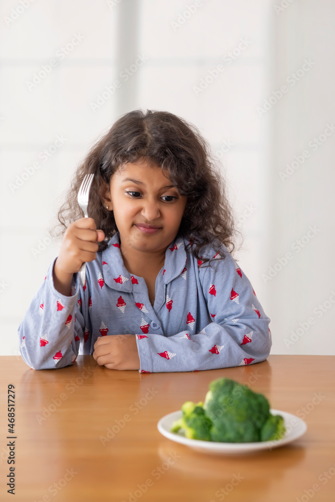 A Girl sitting in front of a plate of broccoli with a disgusted look on her face