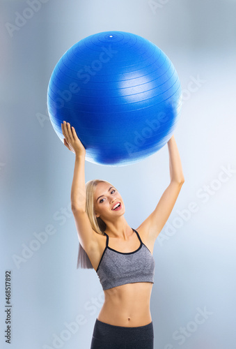 Portrait of happy excited blond young woman in sportswear with fitball, standing over blurred modern gym center background. Fitness, exercising and fit gym concept.