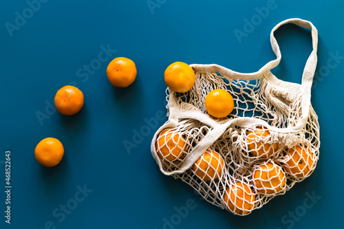 Fresh orange tangerines fruits in cotton eco-friendly mesh bag on blue background.