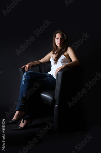 Full length portrait of a beautiful brunette woman sitting on black armchair in front of dark background