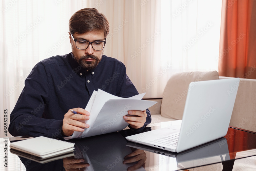 Entrepreneur in eyeglasses works with a laptop and keeps a document in a home office. Man holding paper documents, chatting online with clients on laptop at workplace. Entrepreneur in eyeglasses works with a laptop and keeps a document in a home office. Man holding paper documents, chatting online with clients on laptop at workplace.