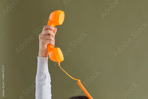 Human hand holding bright vivid orange landline phone receiver. Cropped shot of young lady raising up retro telephone receiver against khaki green blank copy space background. Customer support concept photo