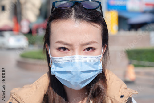 Close up portrait young Chinese woman on street wears protective facial mask looking at camera. Viral flu and coronavirus prevention during outbreak in China and lockdown. Going out during sunny day