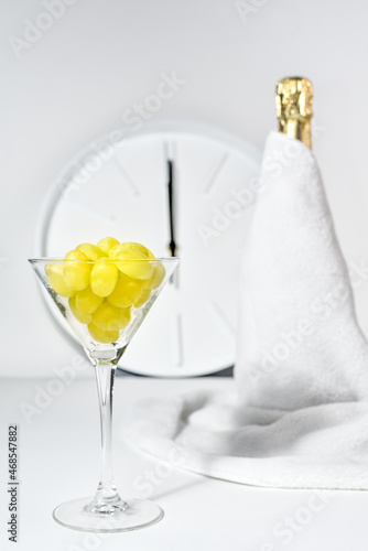 Twelve grapes in a wine glass against the background of a blurry clock and a bottle of champagne. Spanish tradition of eating twelve grapes to celebrate New Year.