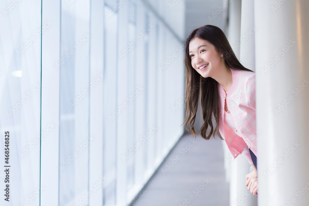 Beautiful teenage Asian girl wears a white with pink shirt and smiles cheerfully look outside while she stands near the glassed window on a nice day.