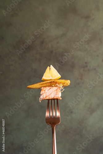 Close-up slice of cooked salmon steak on a fork with mini corn, lemon and microgreens on green background. One slice of grilled salmon on a fork with vegetables