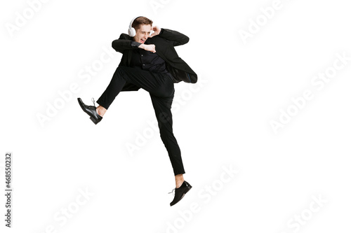Excited young man in black business suit dancing and listening music isolated on white background. Art, motion, action