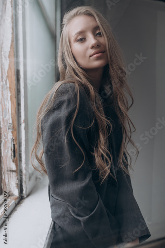 a beautiful girl in a gray jacket with blonde hair is standing by a large window
