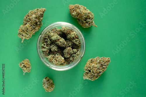 Close up of cbd cannabis buds in glass jar, macro view. Medicinal marijuana blooms on emerald green paper background. Hemp drying flowers, top view.