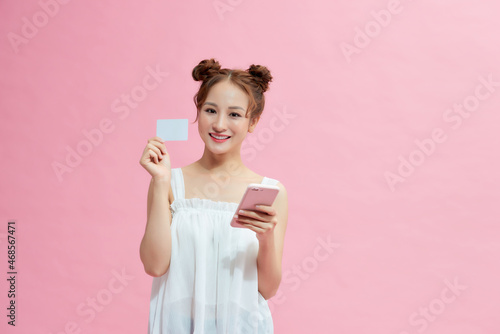Beautiful happy young woman isolated over pink background, showing plastic credit card while using mobile phone